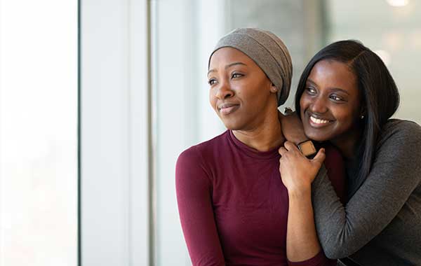 a woman of color with headwrap looks out window with a friend - cancer survivors still need care and support